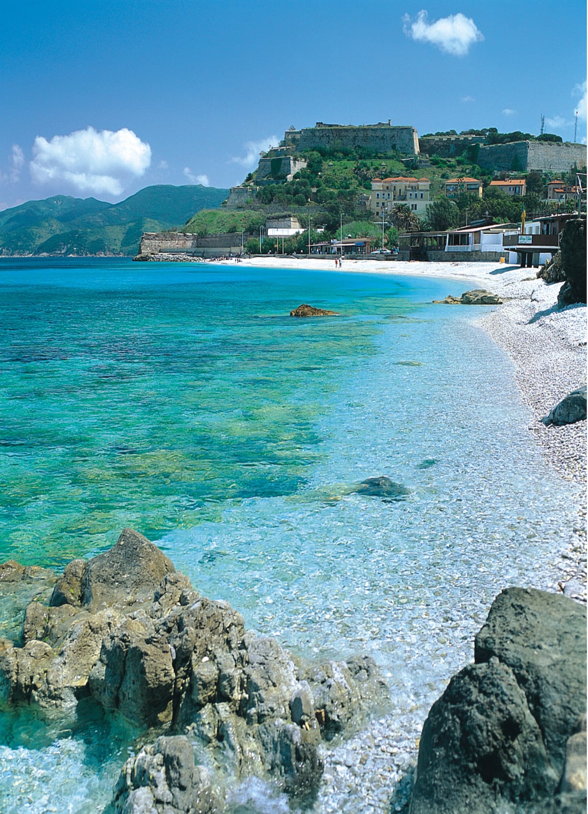 A serene beach scene displays clear turquoise waters gently lapping against a pebbled shore. In the distance, a historic fortress is visible atop a rocky hill, surrounded by a picturesque coastline. Lush green mountains frame the background under a bright blue sky.