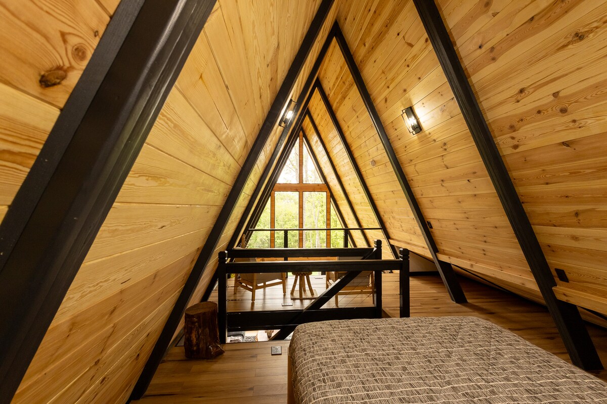 A cozy loft area within the A-frame cabin showcases wooden beams and walls, creating a warm ambiance. A queen-sized floating bed is positioned in the foreground, while a seating area with chairs and a table is visible near the large window, allowing natural light to fill the space.