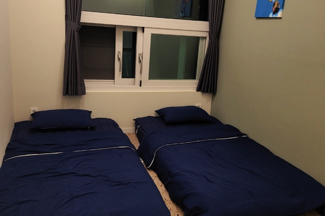 Two spacious sleeping areas are furnished with neatly arranged blue bedding. Natural light enters through a large window adorned with dark curtains. The walls are painted a soft green, creating a calming environment.