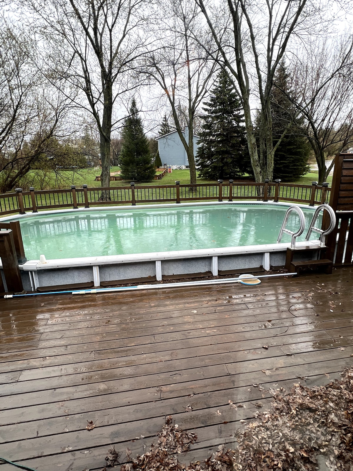 An outdoor above ground pool surrounded by a wooden deck features a set of stairs for easy access. Lush trees and open space are visible in the background, creating a serene setting. The area is designed for relaxation and water enjoyment.