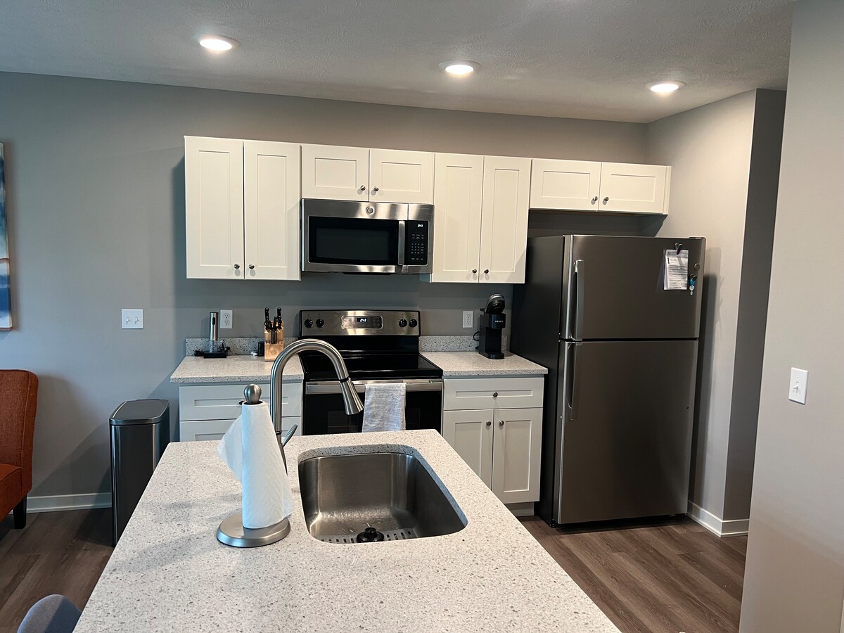 A modern kitchen is displayed with white cabinetry and sleek stainless steel appliances, including a refrigerator and microwave. A spacious countertop with a sink is visible, accompanied by various kitchen appliances. Recessed lighting enhances the bright ambiance in the open space.