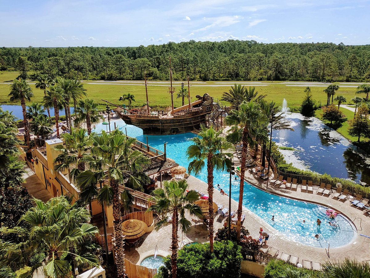 A vibrant pirate-themed pool area is visible, featuring a large ship play structure. Surrounding palm trees provide shade, and sunbeds are arranged along the curved poolside. A serene landscape of green trees and water can be seen in the background, enhancing the outdoor experience.