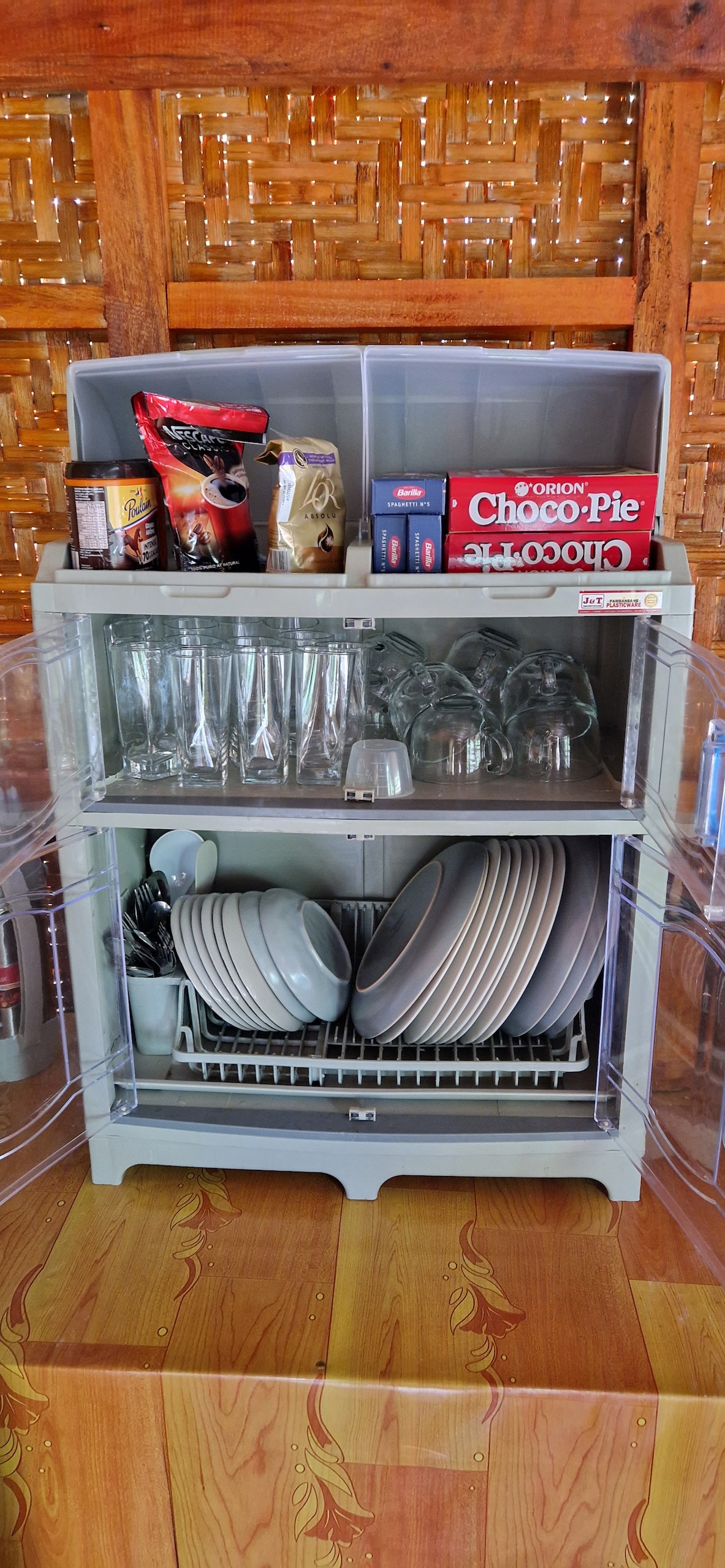 A practical storage unit is shown, featuring a variety of kitchen items. On the top shelf, snacks and coffee products are organized alongside glasses. The middle shelf contains glassware and a drying rack, while the bottom shelf holds a collection of plates, neatly arranged.