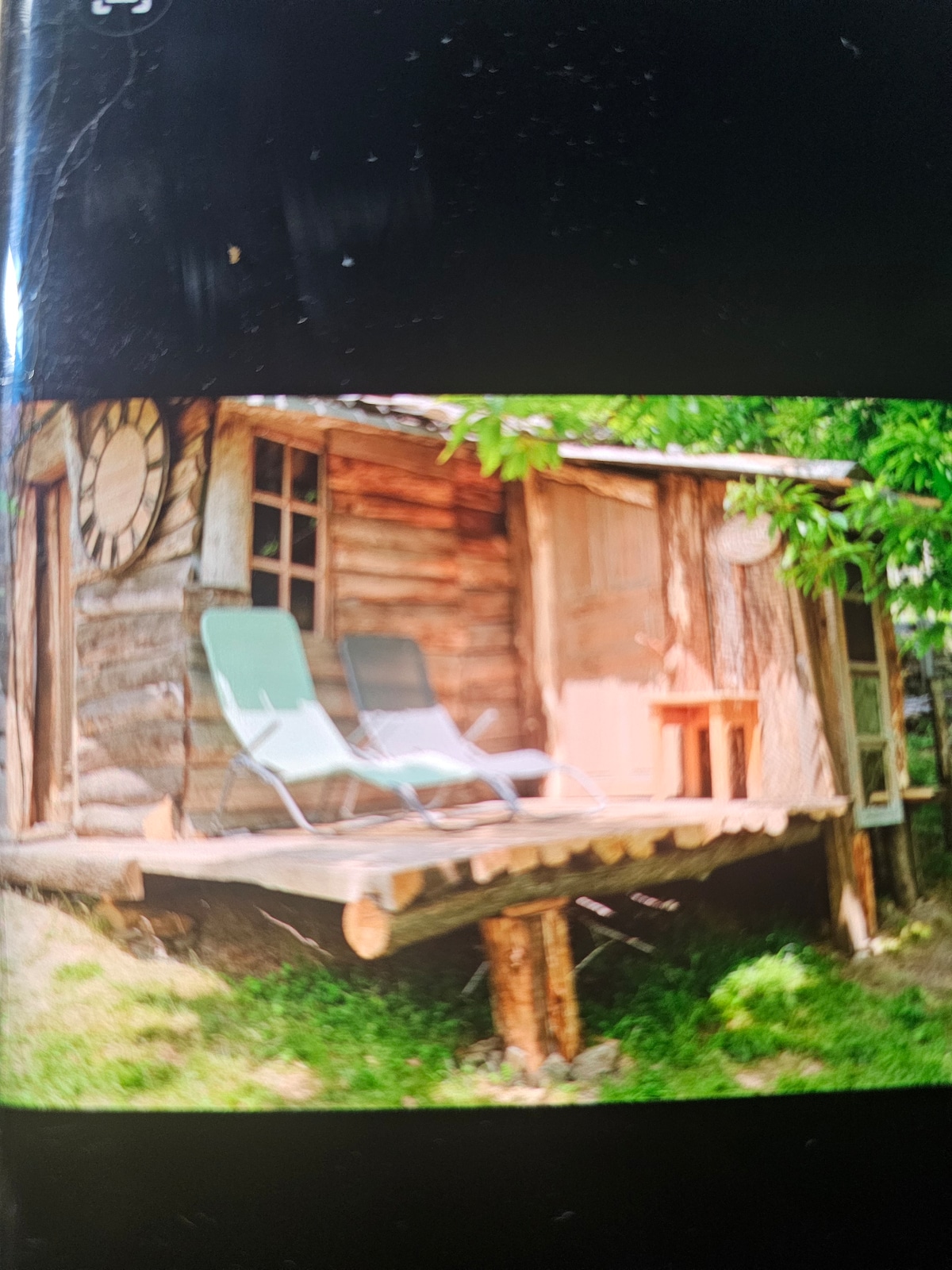 A simple wooden cabin is surrounded by greenery, featuring a small porch with two lounge chairs. The rustic structure has an earthy tone, and a clock is mounted on the wall. Sunlight filters through the trees, enhancing the peaceful outdoor setting.
