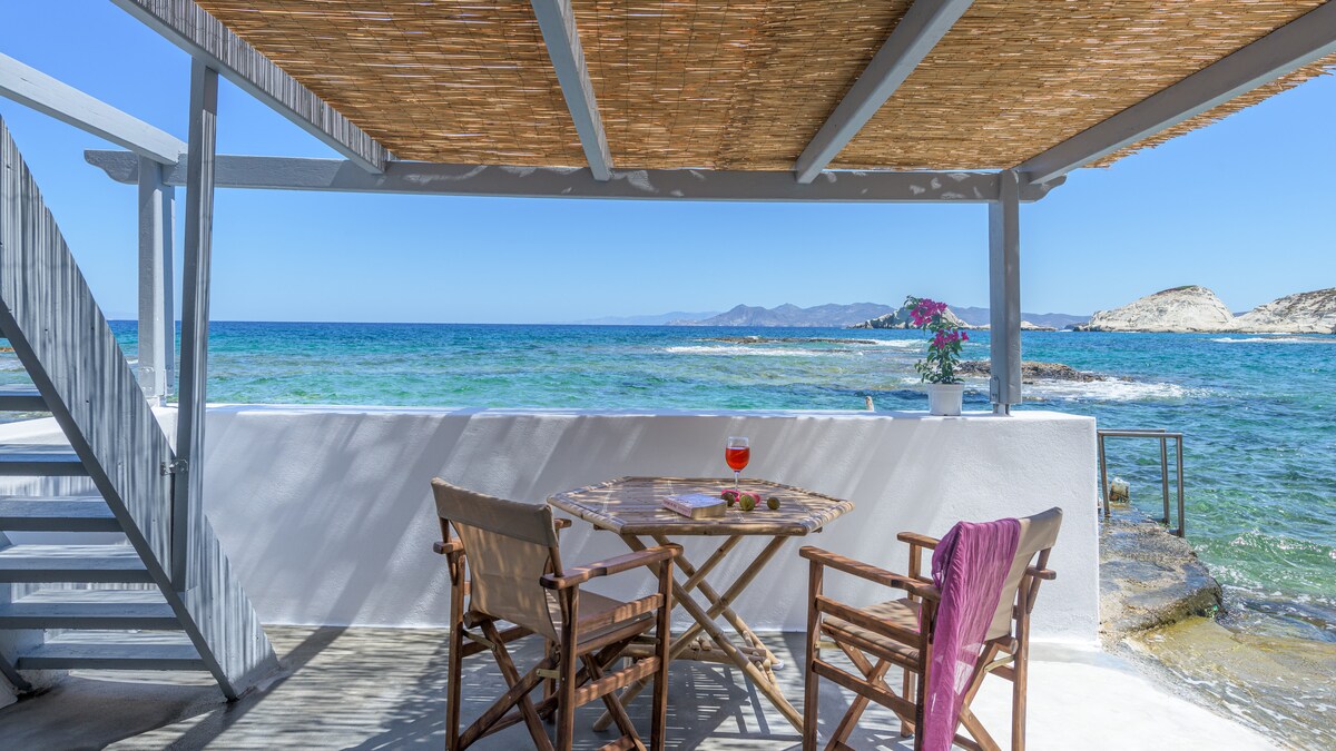 A shaded terrace overlooks the calm sea, featuring a small round table with two wooden chairs. Fresh flowers add a natural touch to the setting. The clear blue waters extend to distant mountains along the horizon, creating a serene coastal ambiance.