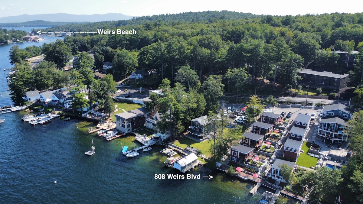 An aerial view captures the waterfront location, showcasing the resort's layout along Lake Winnipesaukee. Nearby, the picturesque landscape features boat slips and access to Weirs Beach. The arrangement of cottages is visible amidst lush greenery and the tranquil lake, enhancing the appeal of the area.
