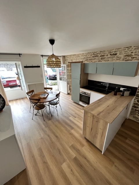 An open kitchen and dining area are featured, with wooden flooring and stone walls adding character. A round table with four chairs is situated next to a large window that allows natural light to enter. The kitchen includes modern appliances on a stylish countertop.
