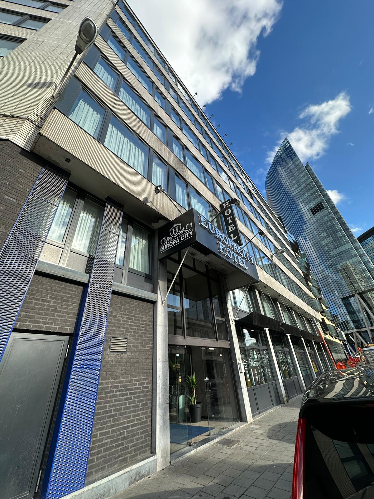 The exterior of a contemporary hotel building is visible, featuring large glass windows and a sleek entrance. The facade combines dark brick with modern design elements. Nearby, a tall, curved skyscraper rises against a clear blue sky, creating an urban backdrop.