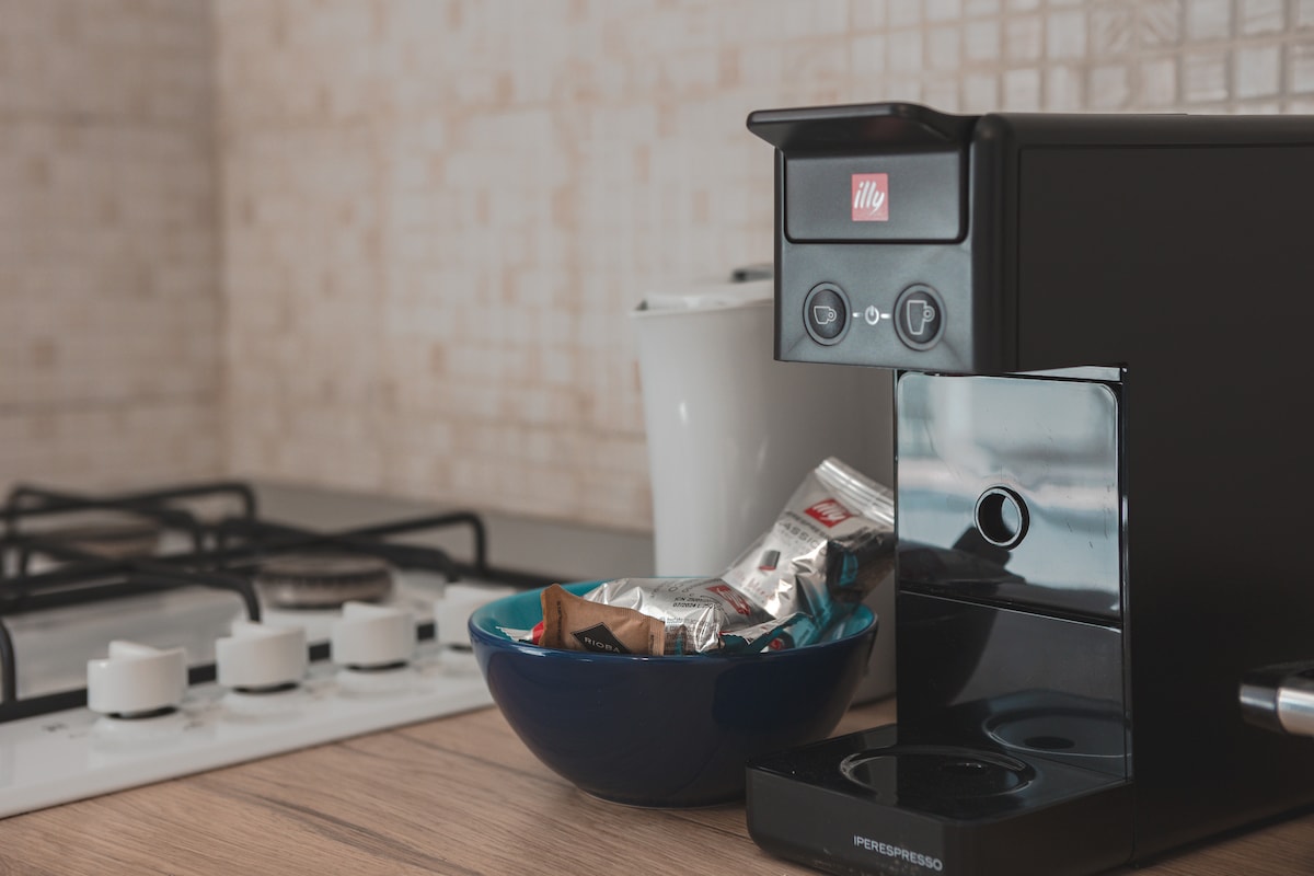 A coffee maker is displayed alongside a selection of coffee pods in a blue bowl on a wooden countertop. White mugs and a gas stove with multiple burners can be seen in the background, creating a functional and inviting kitchen space.