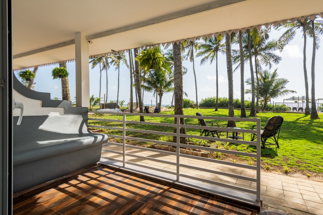A private balcony overlooks a green lawn adorned with palm trees and loungers. The view extends to the beach and sea in the distance. The flooring showcases warm wooden tones, complementing the serene outdoor space.