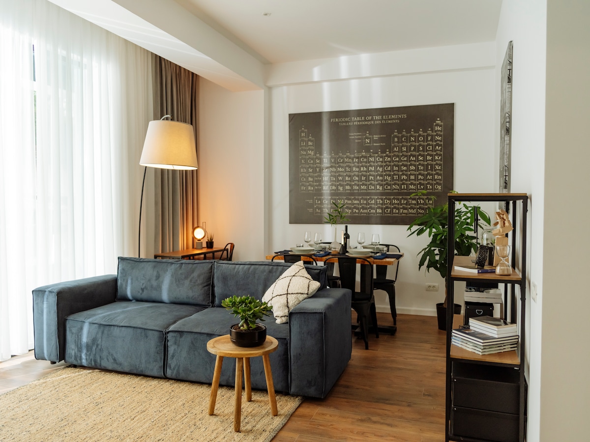 A spacious living area features a modern, sectional sofa complemented by a round wooden coffee table. A large lamp stands beside the sofa, providing soft lighting. A dining table with chairs is visible in the background, along with decorative plants and a framed wall art piece.