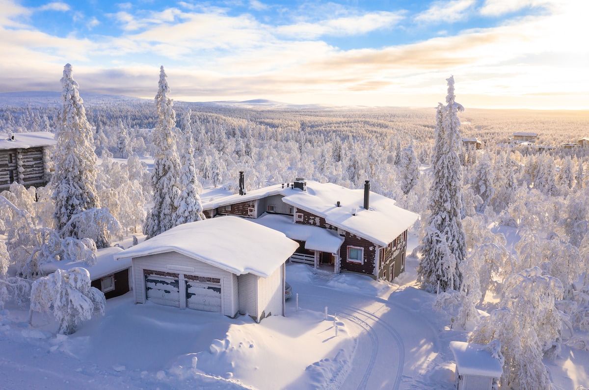 A spacious lodge is nestled in a snowy landscape, surrounded by frost-covered trees. The building features a mix of traditional wooden architecture and modern touches. Soft sunlight bathes the scene, highlighting the serene setting and expansive views of the snow-covered terrain.