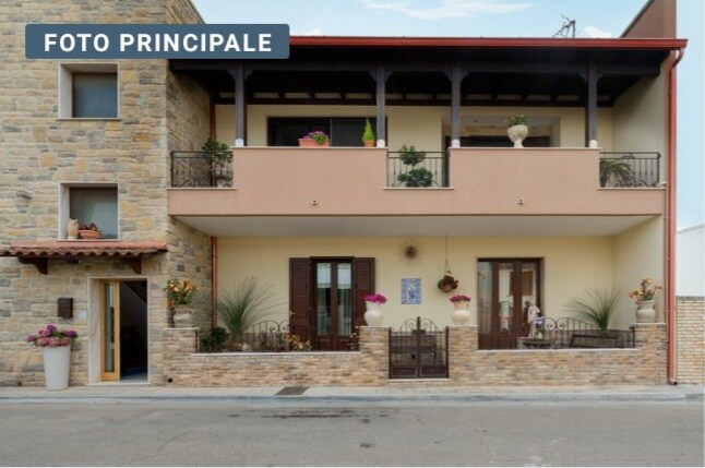 A charming facade showcases a two-story building with a stone exterior and a welcoming entrance. The covered balcony is adorned with potted plants, while the ground level features an inviting terrace surrounded by greenery and ornamental flowers.