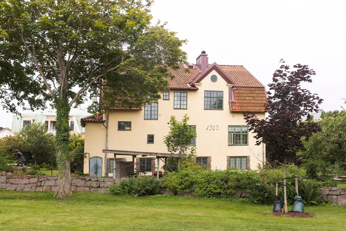 The exterior of Villa Wäring is visible, showcasing a historical building with a beige façade and a distinctive red-tiled roof. Lush greenery surrounds the structure, including trees and shrubbery. The year '1913' is prominently displayed, emphasizing the building's historical significance.