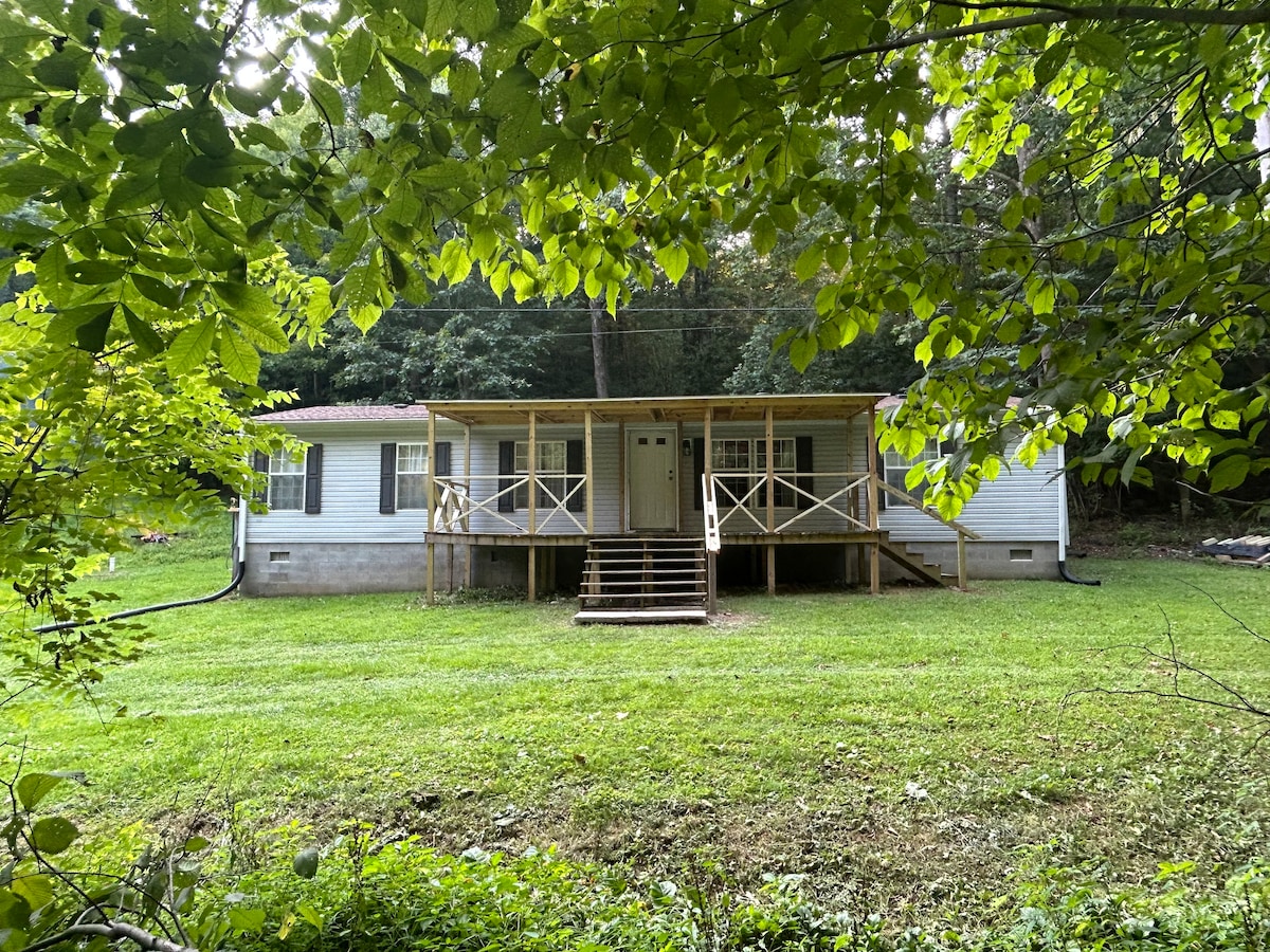 A simple, single-story home is surrounded by lush greenery, featuring a wooden porch with steps leading up to the entrance. The front yard is grassy, providing a natural setting that enhances the home's inviting exterior.