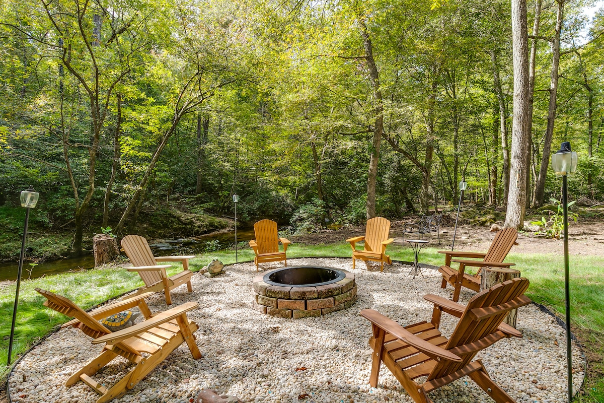 A circular stone fire pit surrounded by six wooden Adirondack chairs is situated on a bed of smooth gravel. Lush greenery and tall trees frame the scene, offering a view of a gently flowing stream in the background.