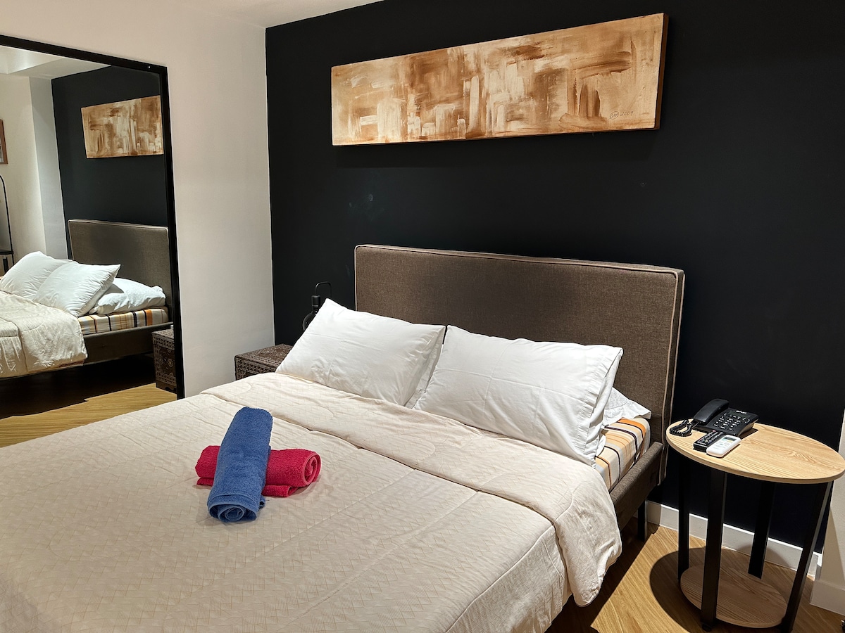 A spacious sleeping area is presented with a neatly made bed featuring white linens and two accent pillows. A mirror reflects the room, while a wooden art piece adorns the dark wall above the bed. A small bedside table holds rolled towels in blue and pink.