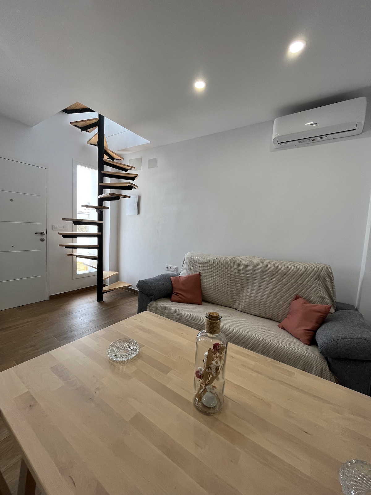 A contemporary interior features a spiral staircase leading to an upper level, complemented by soft lighting. A light wooden table sits in the foreground, accompanied by a cozy sofa with decorative cushions. A glass vase adds a subtle touch to the simple decor.