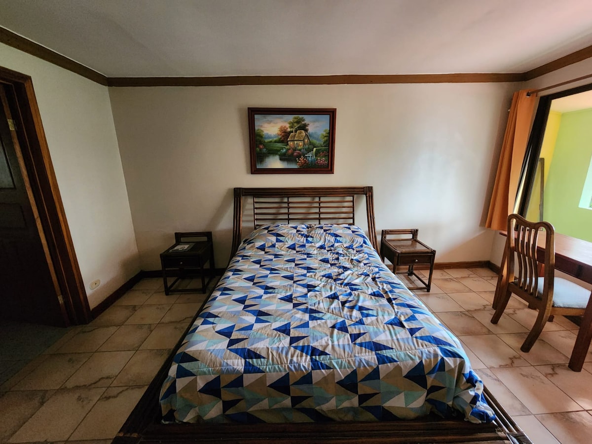 A sleeping area features a spacious bed with a patterned duvet cover. Two wooden nightstands are positioned on either side of the bed. A wooden table is visible near the window, which allows natural light to brighten the room. A framed painting is displayed on the wall.