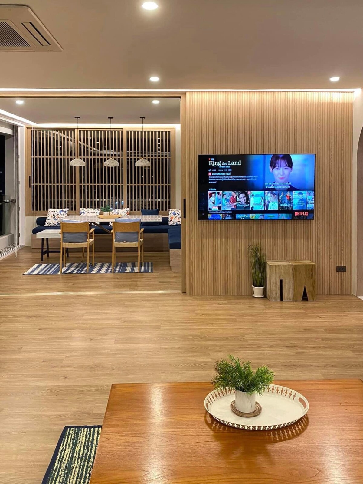 An open living area features a wooden dining table set for six against a backdrop of wooden panels. A wall-mounted television displays content, while a cozy seating arrangement is positioned nearby, complemented by a decorative table and a small plant.