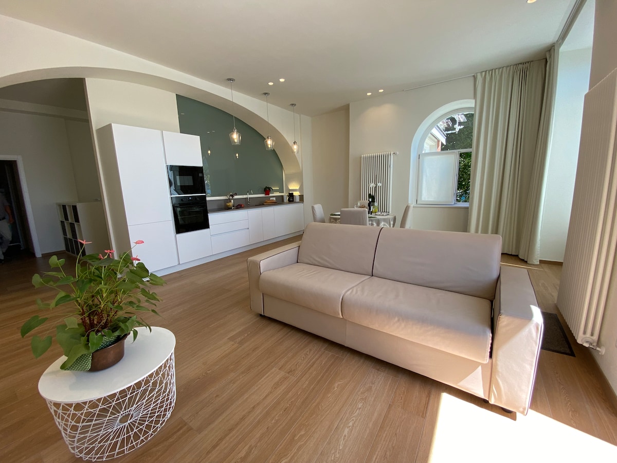 A spacious living area features a light-colored sofa and a round coffee table with a potted plant. The open-plan kitchen boasts modern appliances, while a dining area complements the warm wood flooring. Natural light fills the room through a large arched window, enhancing the bright atmosphere.