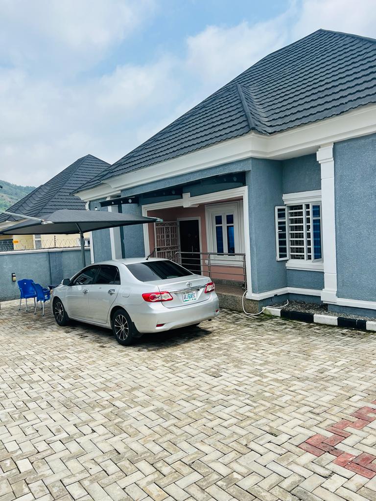 The exterior of the house features a paved driveway with space for a car. A silver vehicle is parked nearby, and a small seating area with two blue chairs is positioned to the side. The house showcases a modern design with a pitched roof and decorative elements.