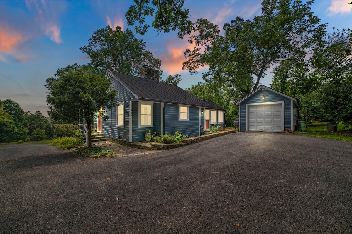 A charming cottage with a blue exterior stands amidst greenery, featuring a welcoming front porch. A garage is visible to the right, and a paved driveway leads up to the home. The sky showcases soft pastel colors, indicating a beautiful sunset.
