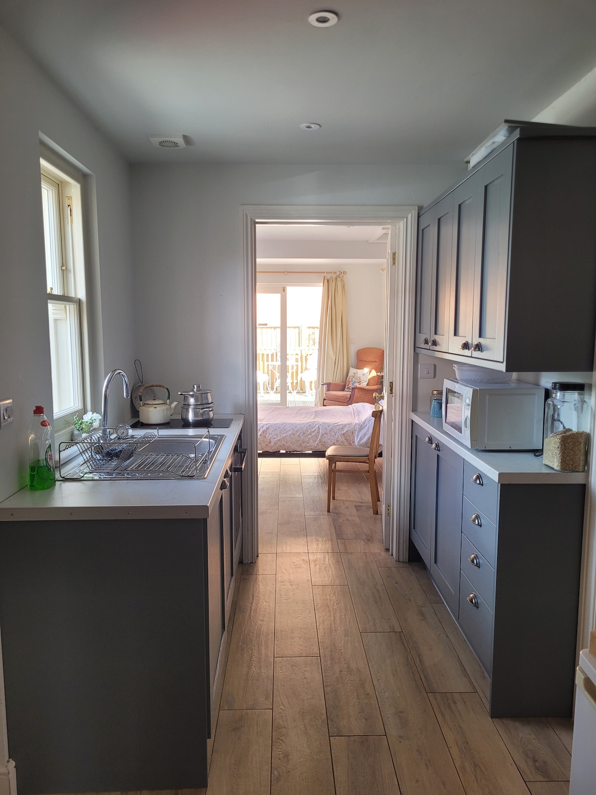 A compact kitchen area features modern cabinetry in gray, with a sink and stove on one side. A light-colored countertop provides space for appliances, including a microwave. The open doorway leads to a bright bedsitting room, illuminated by natural light.