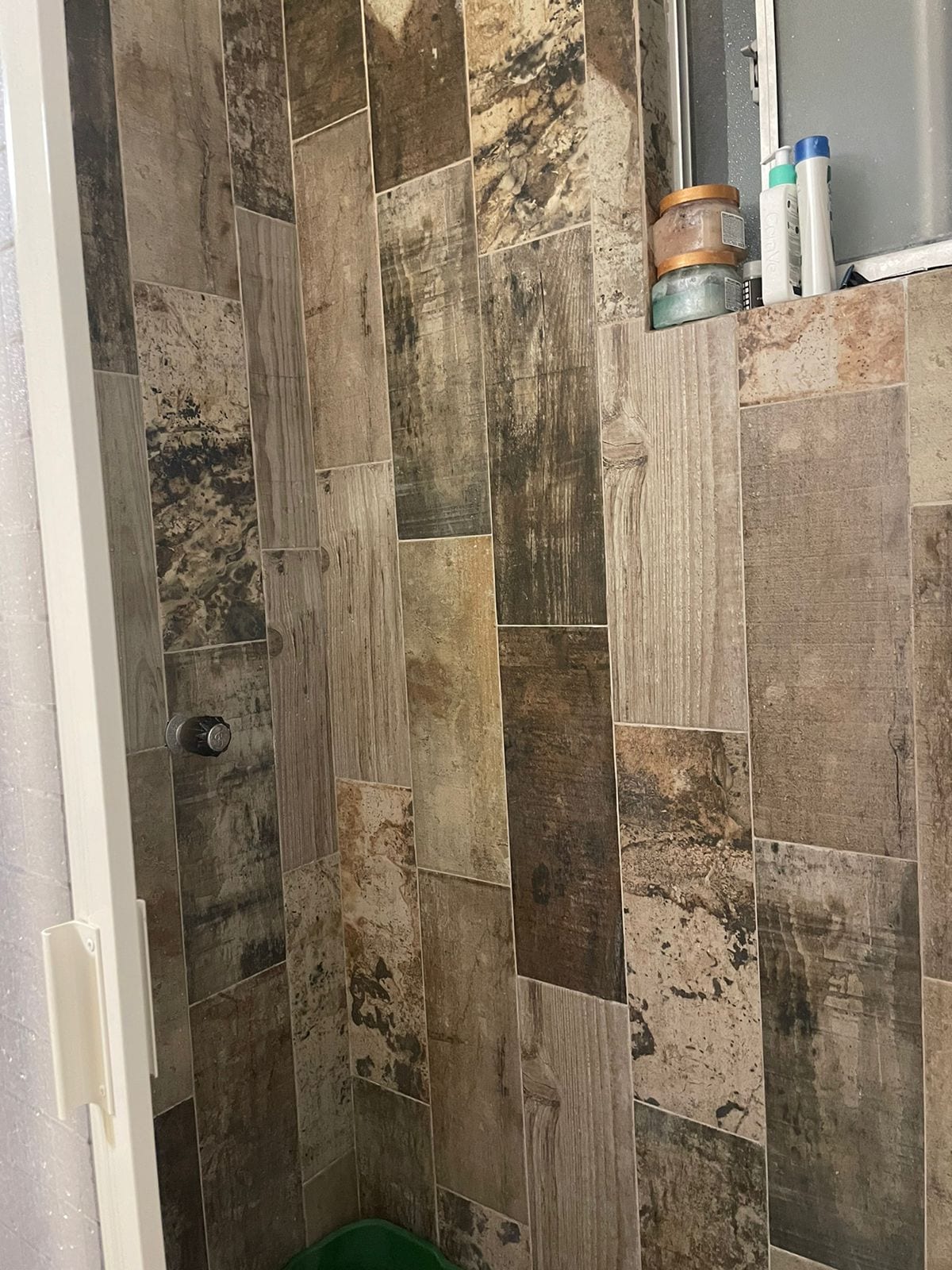 A modern shower space features textured tiles in neutral tones, arranged in a vertical pattern. Shelves hold personal care items, while a showerhead is mounted on the wall. Natural light enters through a small window, enhancing the area’s functionality.