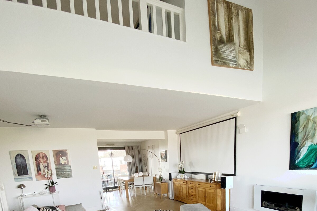 The spacious living area features high ceilings and a mezzanine level, with a large, minimalist entertainment unit beneath a wall-mounted screen. Modern art adorns the walls, and natural light fills the room through large windows that open to a terrace.