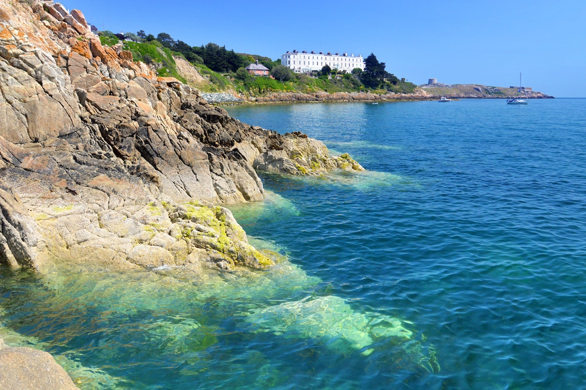 Clear turquoise waters meet rocky shoreline, showcasing vibrant marine life among the rocks. The distant white buildings contrast with the natural scenery, while a few boats are visible on the calm water, indicating a tranquil coastal setting.