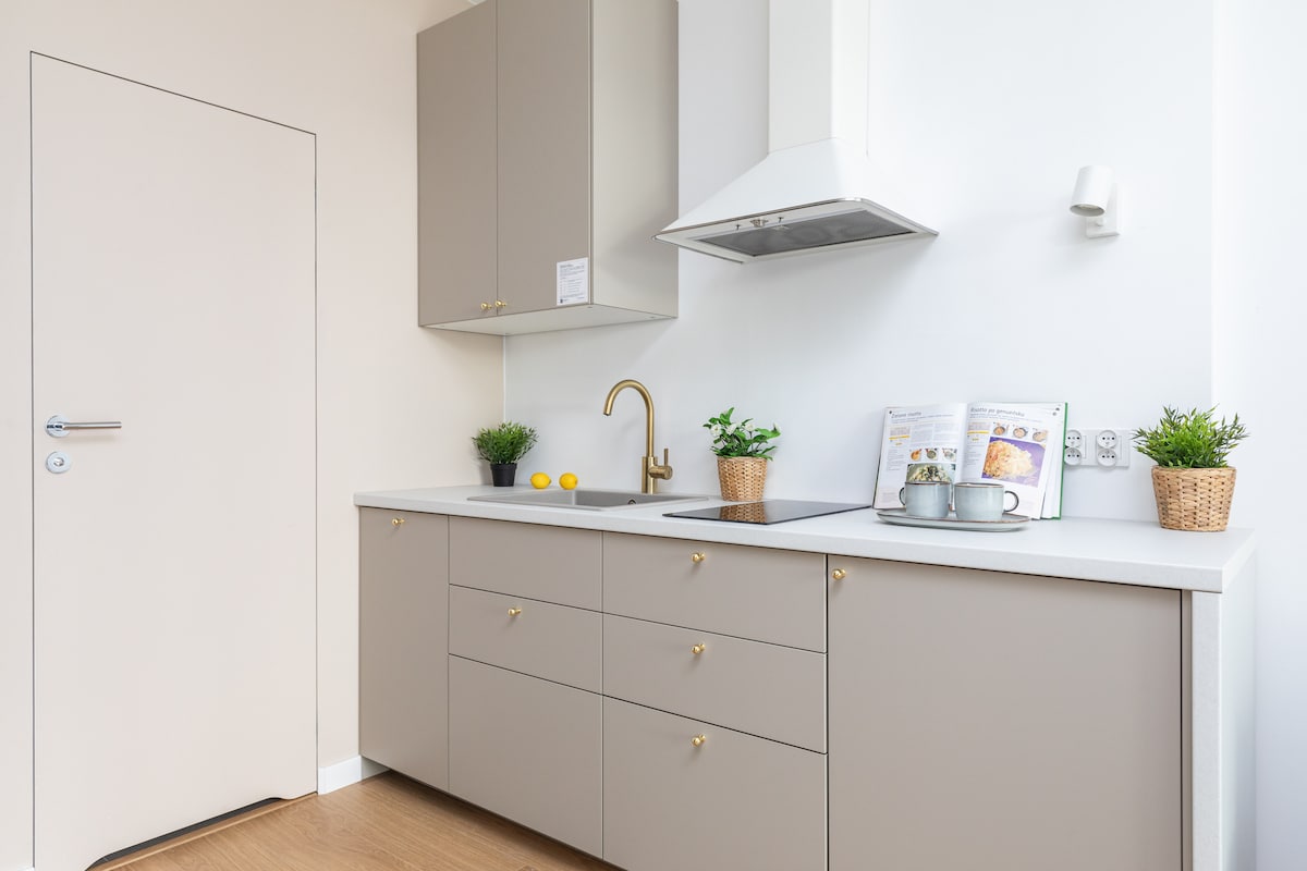 A modern kitchen area is equipped with grey cabinetry and a white countertop. A sink with a gold faucet is positioned under a wall-mounted range hood. Small potted plants and a cookbook are displayed on the counter, enhancing the functional and inviting space.