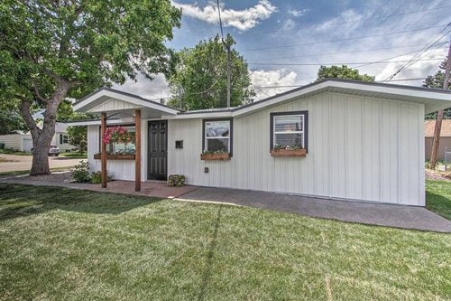 The exterior of the cottage showcases clean lines and a modern design. Flower boxes adorn the windows, adding a touch of color. A cozy front porch is supported by wooden pillars, leading to the entrance, surrounded by well-maintained grass and trees that provide shade.