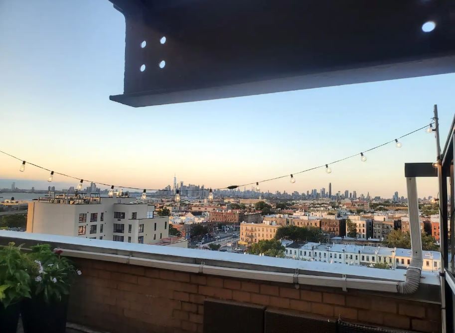A private roof deck offers panoramic views of the city skyline, with illuminated string lights above. The scene includes various rooftops and greenery, creating an inviting space for relaxation or gatherings.