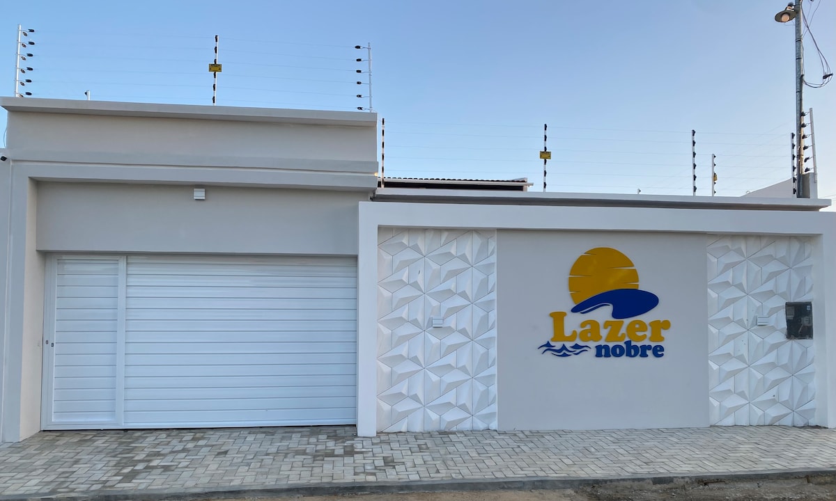 The exterior of the home features a modern facade with a smooth white finish and a geometric design on the wall. A prominent logo displaying 'Lazer Nobre' is positioned under a sun graphic. The driveway consists of paved stones, providing an inviting entrance.
