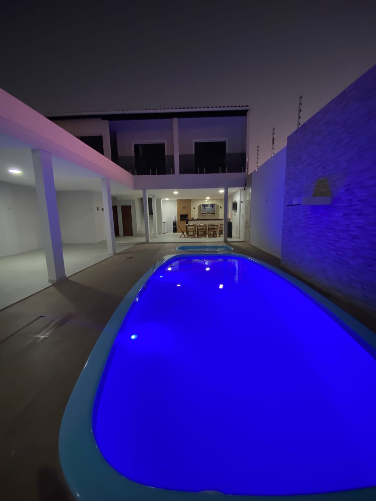 A serene pool area is illuminated in calming blue tones, surrounded by sleek modern walls. A spacious covered seating area is visible in the background, along with an open kitchen space, creating an inviting setting for relaxation and gatherings.