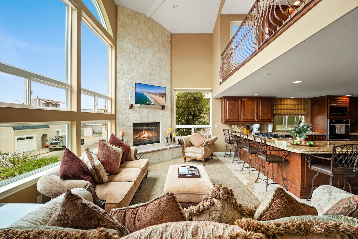 A spacious living room features large windows allowing natural light to fill the area. Cozy seating arrangements include a sectional sofa and armchair. A fireplace is centrally positioned, with a large TV mounted above it. The open layout connects to a well-equipped kitchen with seating at the bar.