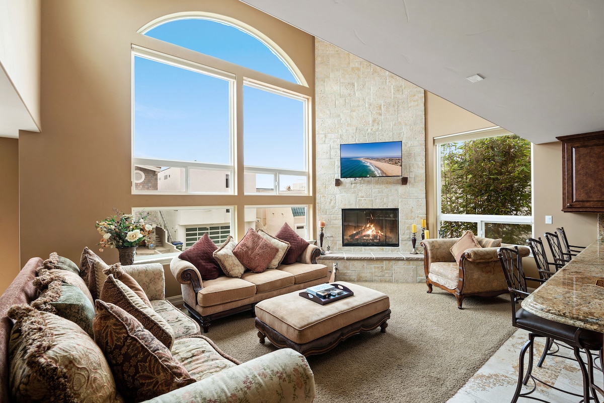 The living room showcases a welcoming arrangement of two couches and an armchair, harmonizing with a stone fireplace accented by a mounted TV. Natural light spills through large windows, illuminating the cozy space, while a stylish coffee table is positioned atop a plush area rug.