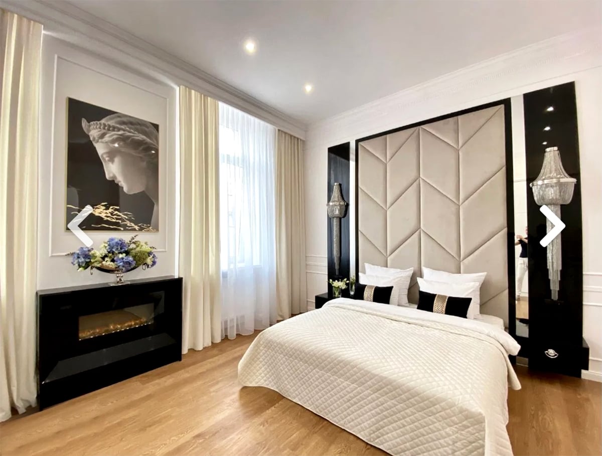 The sleeping area features a spacious bed with a neutral-coloured coverlet. Flanking the bed are two elegant bedside tables with lamps. A decorative wall art piece is displayed above a sleek fireplace, while sheer curtains frame a large window, allowing natural light to fill the room.
