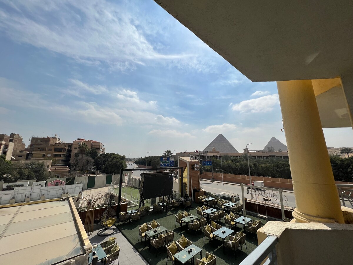 A balcony view showcases the iconic pyramids in the distance, framed by a clear blue sky. Below, an outdoor seating area features numerous tables and chairs, surrounded by greenery, creating an inviting space for relaxation.