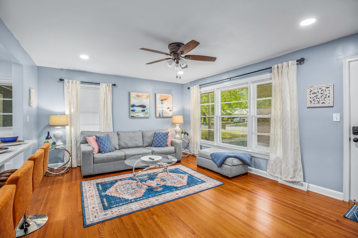 A bright living room features a comfortable gray sofa with colorful accent pillows, set on a decorative area rug. Large windows provide natural light, and light curtains frame the view. A ceiling fan and art pieces enhance the space, alongside a small decorative table.