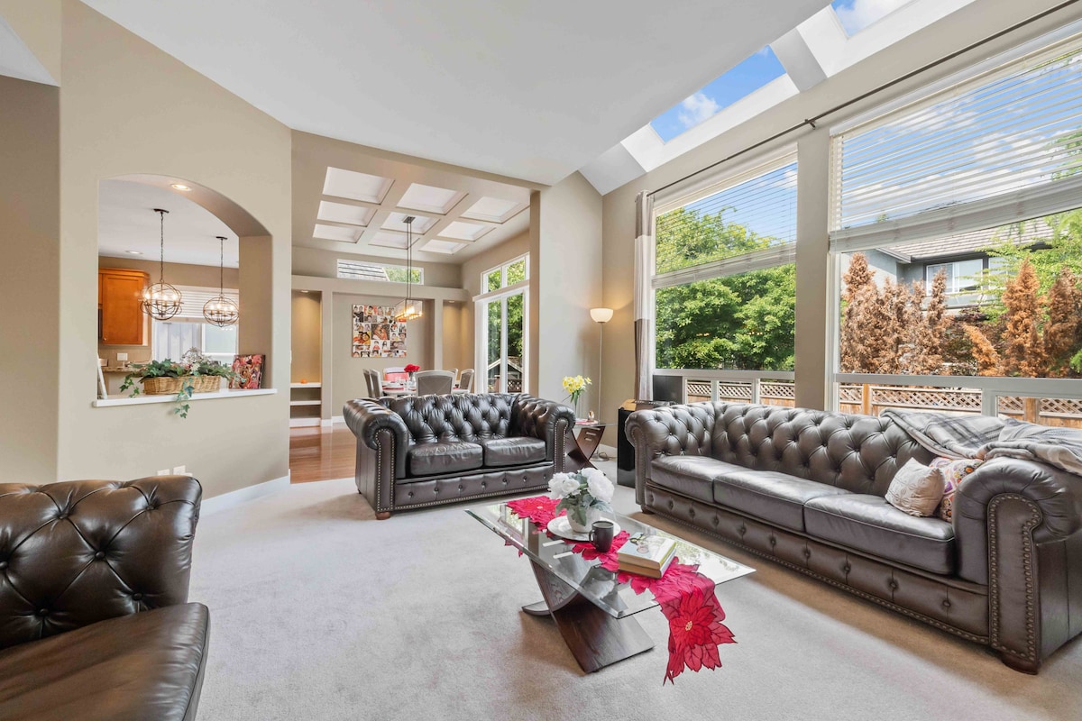 A bright living room features large windows allowing natural light to illuminate a cozy seating arrangement. Two dark leather sofas face a glass coffee table, adorned with a decorative arrangement. The open layout leads towards a dining area visible in the background.