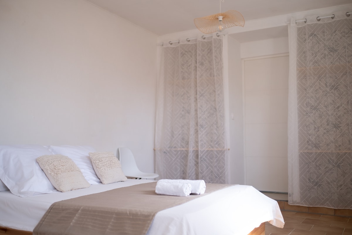A comfortable sleeping area is presented with a spacious bed featuring light linens and accent pillows. Soft natural light enters through sheer curtains, creating a peaceful ambiance. A white chair is positioned nearby, adding a minimalist touch to the room's design.