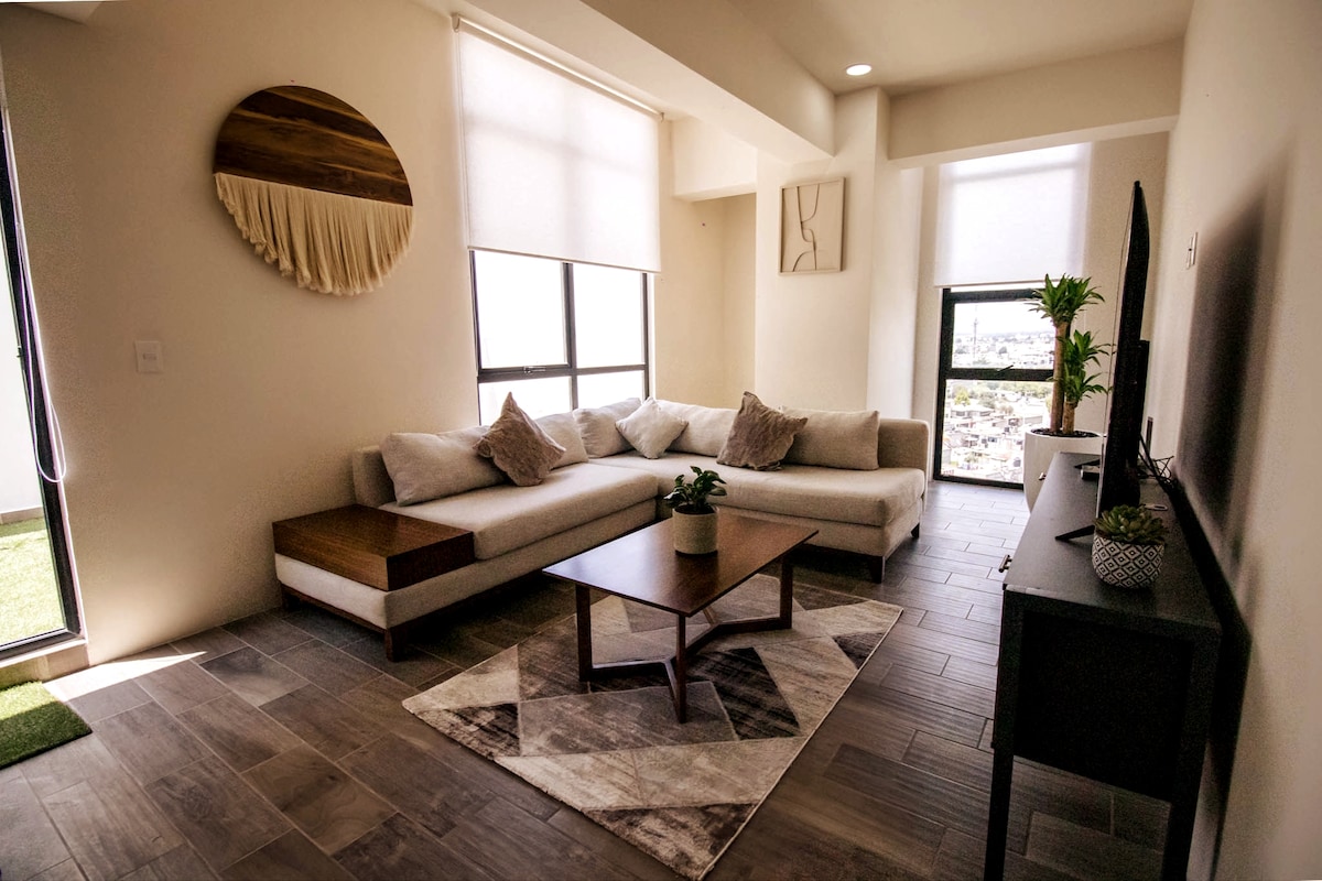 A spacious living area features a comfortable sectional couch, a coffee table on a geometric rug, and a sleek entertainment center with a flat-screen TV. Large windows provide natural light, and a small potted plant adds a touch of greenery.