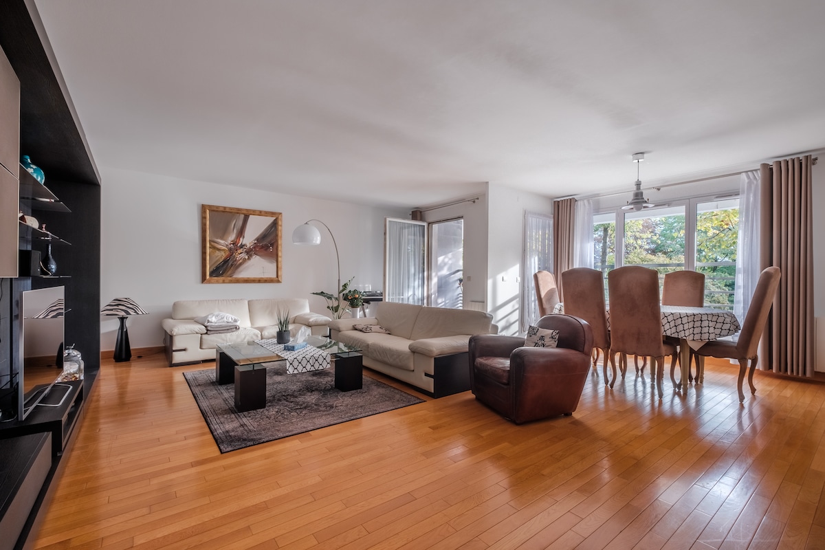 A spacious living area features large windows allowing natural light to fill the room, showcasing wooden flooring. Two comfortable sofas in neutral tones are complemented by a glass coffee table. A dining area with a large table and supportive chairs occupies one side, creating an inviting space.