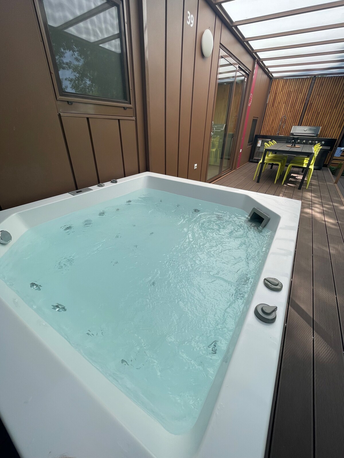 A spacious outdoor area features a modern Jacuzzi surrounded by wooden decking. Nearby is a dining table with chairs, offering a delightful spot for relaxation and social gatherings. Natural light filters through a transparent roof, enhancing the inviting atmosphere.