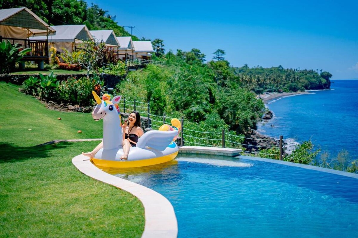 A vibrant inflatable unicorn floats in an infinity pool, surrounded by lush green grass. Beachfront tents are visible in the background, along with the clear blue sea and a rocky shoreline. Sunlight illuminates the scene, enhancing the pool's inviting appearance.