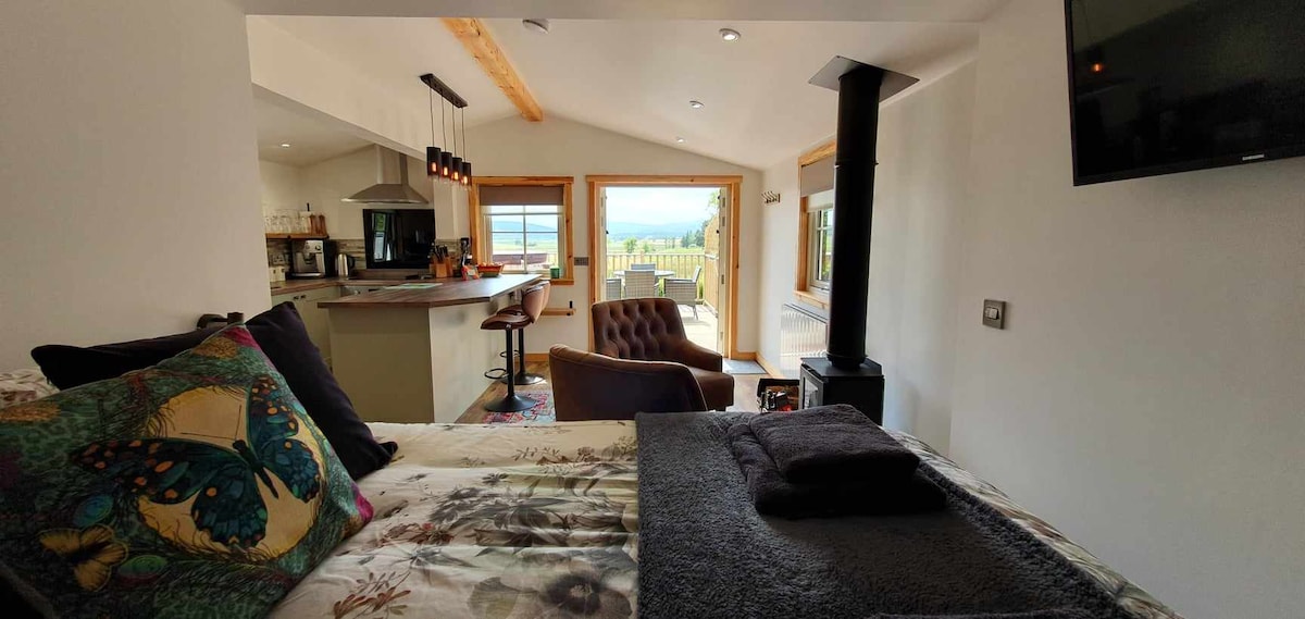 An open-plan living area features a comfortable bed with decorative pillows, a cozy chair, and a woodburning stove. Large glass doors provide access to a raised deck, offering views of the surrounding landscape and allowing natural light to fill the space.
