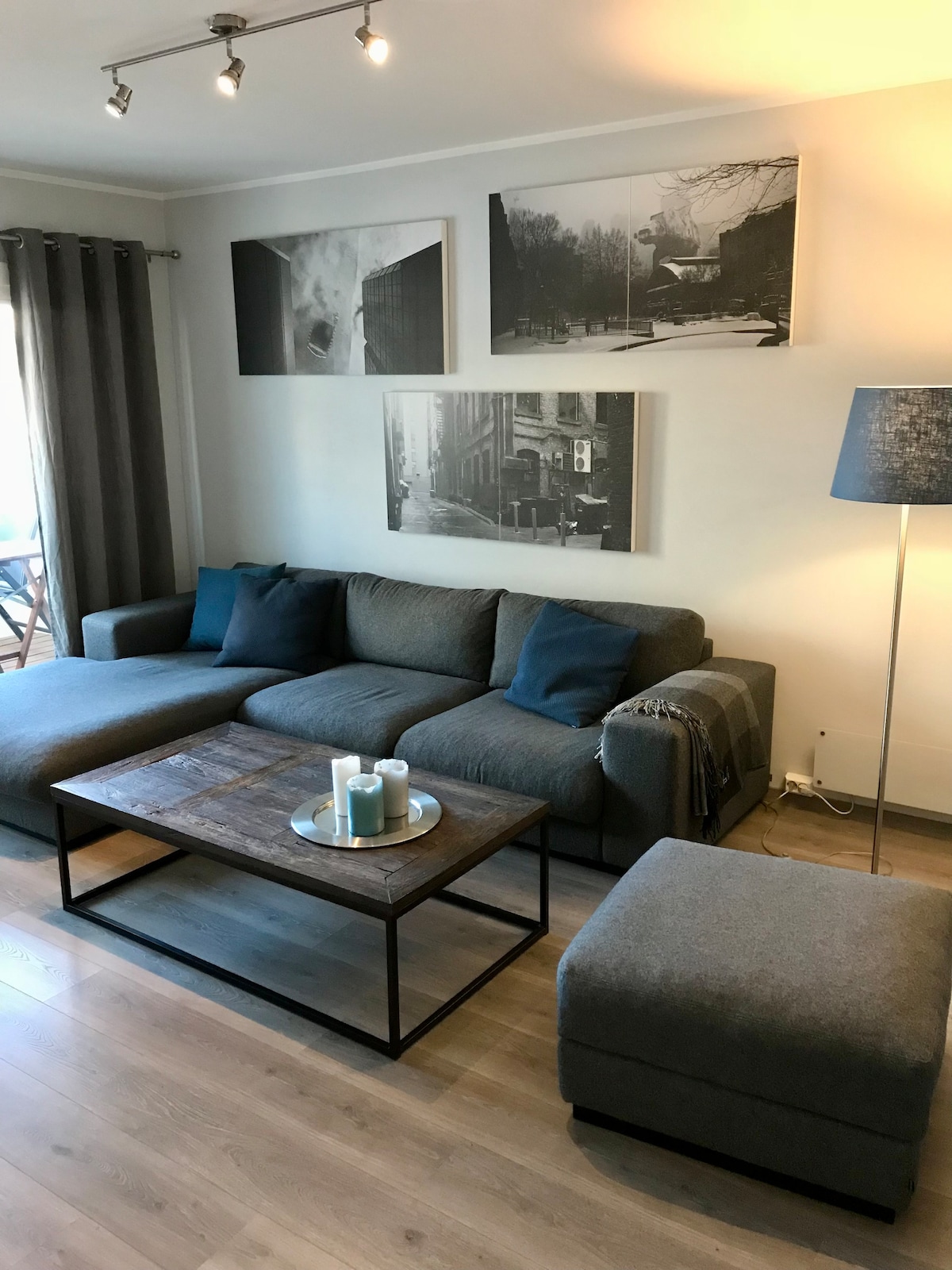 A comfortable living area features a large gray sectional sofa adorned with accent pillows. A wooden coffee table sits in the center, holding decorative candles. Two framed black-and-white photographs decorate the walls, and a floor lamp stands next to the seating area.