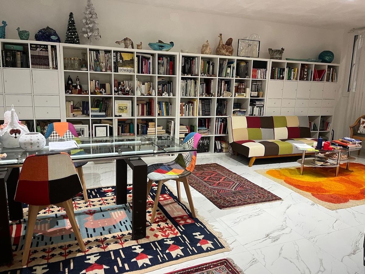 A spacious living area is highlighted by a large glass dining table set with colorful chairs. A modern sofa is positioned nearby, surrounded by an extensive bookshelf filled with books and decorative items. Various vibrant rugs enhance the room's visual appeal, while soft light filters through nearby curtains.